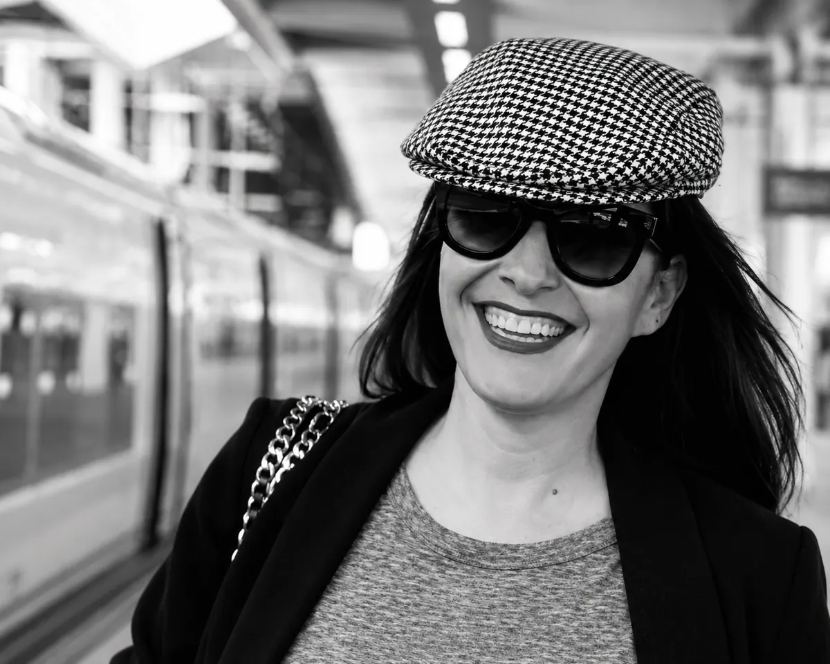 Jessica Bataille smiling on a station platform, wearing a houndstooth cap and dark glasses